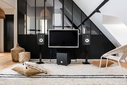 Triangle BR02 BT højttalere i askegrå på tæppe i moderne stue med trappe, glasvæg, designersofa og lænestole