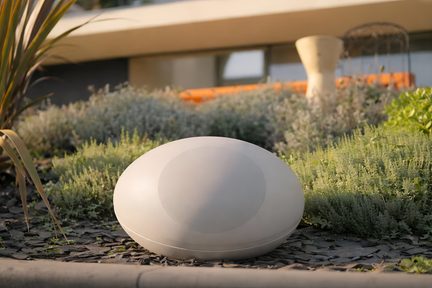 Diffusore da esterno bianco a forma di ciottolo, posato su ardesia in un giardino contemporaneo