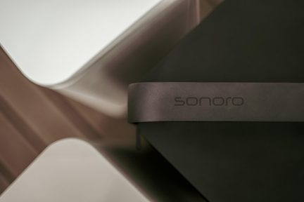 närbild på Sonoro-logotyp på mörkgrå högtalarlist med designutseende, suddig geometrisk beige och grön bakgrund