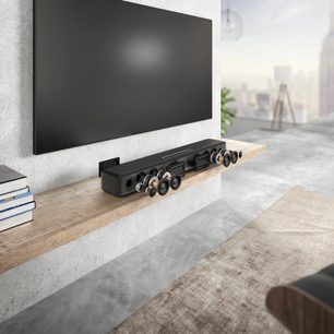 Denon Home 550 black soundbar placed on a light wood shelf in front of a wall-mounted black television screen turned off