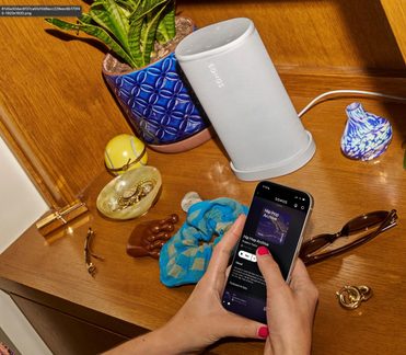 Hvid cylindrisk trådløs Sonos-højttaler på træhylde, med Sonos-appen vist på smartphone i hånden
