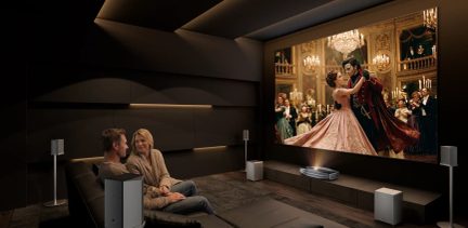 Hjemmekinoprojektor i mørkt rom med gigantisk lerret, par sittende på sofa og høyttalere rundt