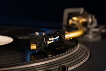 Nærbillede i trekvart vinkel af en sort fonografcelle i metal og plast med synlige kabler, monteret på en gylden metal-tonearm og placeret på en sort vinylplade