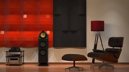 Pohled zepředu na poslechový prostor s hi-fi vybavením, s dřevěnou podlahou, centrálním černým lakovaným sloupovým reproduktorem se žlutým kalotem, kovovým audio rackem vlevo, červenými lesklými a černými matnými čtvercovými akustickými panely v pozadí, lounge křeslem a černou koženou otomanem s dřevěným zakřiveným rámem vpravo, stojací lampou s červeným stínidlem na trojnožce.