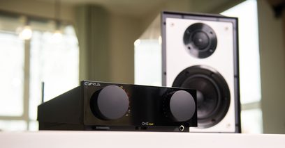 Ampli hi-fi stéréo Cyrus ONE HD Cast noir, vue de face avec deux boutons rotatifs et deux enceintes acoustiques en arrière-plan