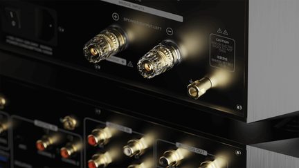 Zlaté audio konektory na zadním panelu zesilovače, pohled ze šikmého úhlu, viditelné bezpečnostní štítky
