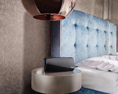 Trådløs tilsluttet Denon HEOS 3 HS2 sort højttaler på rund beige sokkel, foran lys blå polstret sengegavl