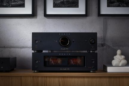 Dva černé hi-fi prvky z kovu, pohled zepředu, předzesilovač s velkým knoflíkem a zesilovač s oranžovými VU metry na dřevěné polici