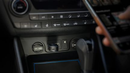 Nahaufnahme, Frontansicht, eines schwarzen, matten Kunststoff-Zylinderadapters mit USB-Anschluss, der in eine 12-V-Steckdose in der Mittelkonsole eines Autos eingesteckt ist, umgeben von ähnlichen Steckdosen und Klimaanlagen-Knöpfen.