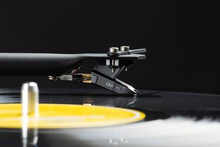 Um braço de toca-discos preto com uma cápsula OM 10 prateada e preta, colocado sobre um disco de vinil com um rótulo amarelo