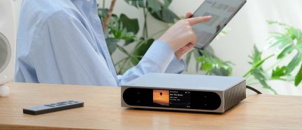 Compatto case audio rettangolare in alluminio argentato spazzolato, vista tre quarti anteriore destra, frontale con schermo centrale a colori incorniciato da superfici touch nere e lato destro con alette di dissipazione