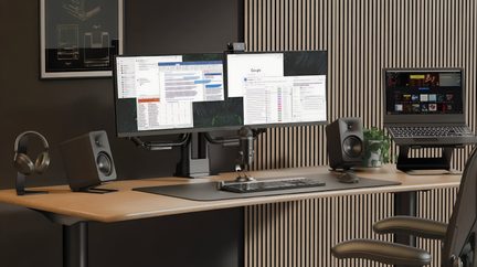 Rechteckiger Schreibtisch aus hellem Holz, Frontansicht, ausgestattet mit zwei schwarzen Flachbildschirmen auf Metallarmen, einem schwarzen Laptop auf einer Halterung, zwei schwarzen Desktop-Lautsprechern, einem zylindrischen Mikrofon aus gebürstetem Silbermetall, einer schwarzen Tastatur und Maus, einem schwarzen Kopfhörer auf einem Metallständer sowie einer grünen Topfpflanze, an eine Wand mit vertikalen Lamellen aus hellem Holz gelehnt