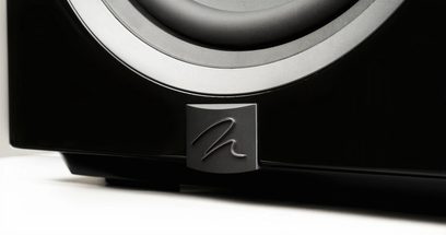 Dynamo 12 subwoofer med sort højglansoverflade, delvis nærbillede i vinkel, metallogo i relief på frontpanelet