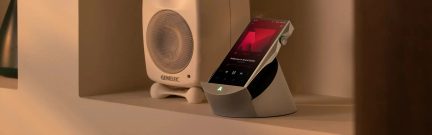 Lettore audio Astell&Kern argentato su supporto, schermo touch inclinato e manopola laterale accanto a un diffusore Genelec bianco