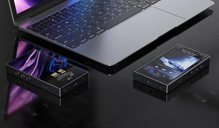 Grå bærbar computer i aluminium og to kompakte sorte metal-lydafspillere med sølvfarvede, affasede kanter, farvedisplay og sideknapper, placeret på en sort, reflekterende overflade set fra let ovenfra