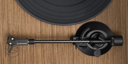 Sort kulfiber-tonearm til vinyl-pladespiller Audio-Technica på rundt sort pladetallerken og sort rund sokkel med lys egetræsfinish