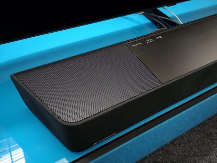 Černý soundbar na modrém povrchu, pohled shora a z boku. Kovový gril na horní části soundbaru