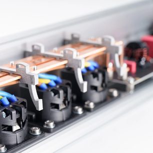 Detailní šikmý záběr interiéru prodlužovacího kabelu Inakustik AC-3200 Filter Analog | SCHUKO, hliníkový stříbrný rám, měděné vodivé lišty upevněné šedými kovovými svorkami, černé kruhové zásuvky a modro-žluté kabely