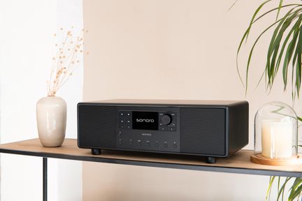Czarny, zintegrowany system audio Sonoro z centralnym ekranem i głośnikami stereo ustawiony na drewnianej półce w minimalistycznej aranżacji