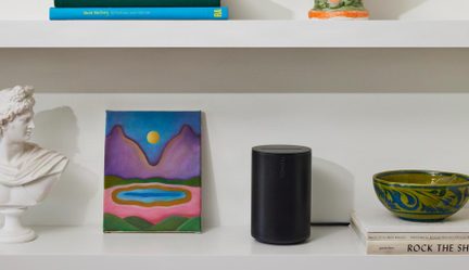 Čierny valcovitý bezdrôtový reproduktor Sonos na bielej polici medzi farebným obrazom a keramickou misou
