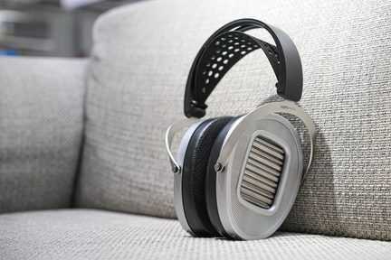 Hovedtelefon i sølvfarvet metal, sort perforeret hovedbøjle, ovale ørepuder med gitter, trekvartsvisning på stofsofa
