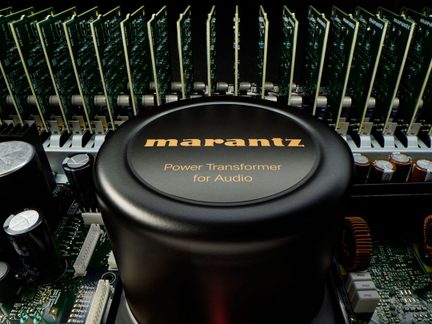 Közeli felülről nézet egy fekete, hengeres, toroidális fémtranszformátorról, amelyen a "marantz Power Transformer for Audio" felirat olvasható, körülötte zöld, függőleges elrendezésű elektronikai lapokkal, kondenzátorokkal és réz tekercsekkel.