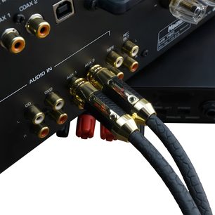 Primo piano, vista posteriore leggermente obliqua, di due cavi audio analogici RCA Oehlbach Black Connection Master 1,25 m con guaina nera testurizzata e connettori cilindrici in metallo dorato collegati alle prese AUX 1 di un amplificatore