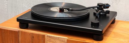 Tocadiscos negro con brazo de lectura, plato circular, disco de vinilo colocado, patas cilíndricas, superficie de madera clara
