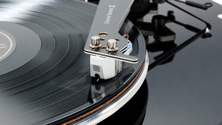 Skråt nærbillede, der viser en sort metal-tonearm med to kromskruer og en sølvgrå pickup med metallisk finish, placeret på en skinnende sort vinylplade, der drejer på en sort pladetallerken med sølvkant