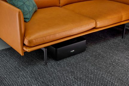 Kompaktowy czarny subwoofer Canton pod camelową skórzaną kanapą na melanżowym szarym dywanie