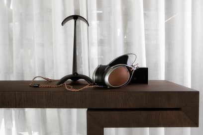 Åben kablet hi-fi-hovedtelefon Meze Audio Poet placeret på et mørkt træbord med sort holder, snoet kabel og hvidt gardin i baggrunden