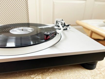 Platine vinyle argentée en métal, plateau aluminium, bras blanc courbé à cellule rouge, disque noir, vue de trois-quarts