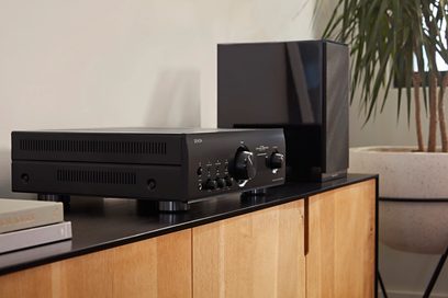 Ampli hi-fi stereo Denon PMA-1700NE černý, pohled zepředu, položený na světlém dřevěném nábytku, se zelenou rostlinou v pozadí