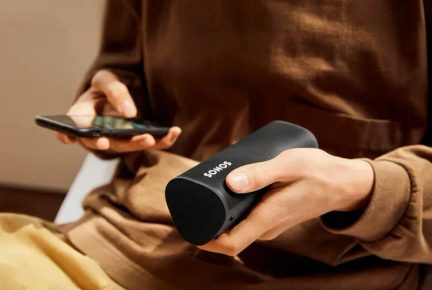 Front view of a black Sonos portable speaker held in hand, with the touch screen illuminated