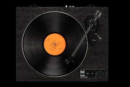 Toca-discos preto com acabamento em madeira escura, prato com disco de vinil preto e etiqueta laranja, braço de leitura metálico inclinado e painel de controle preto exibindo o logotipo Dual no canto inferior direito, visto de cima