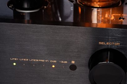 Nærbillede af Cayin CS-805A’s frontpanel i børstet sølvfarvet aluminium med stor sort vælgerknap til højre, central række af grønne og orange LED-indikatorer for indgang, samt orange glød fra rørene synlig i baggrunden