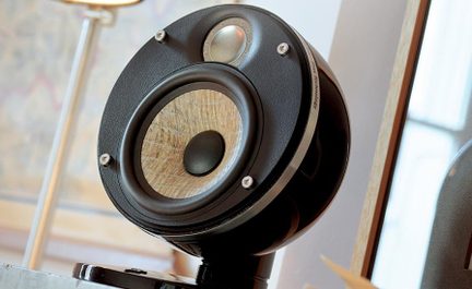 Kompakt højttaler i sortlakeret sfærisk kabinet, set i trekvart vinkel let nedefra, fronten med en beige vævet fiber-keglewoofer omgivet af gummi, en sølvfarvet metal-diskant og fire synlige skruer