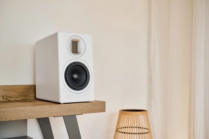 Altavoz de audio blanco rectangular, altavoz negro, rejilla dorada, sobre estante de madera clara, pantalla de mimbre al lado