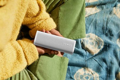 Sonos Roam Lunar White Bluetooth Speaker, White finish, held in one hand against a yellow sweater and faded jeans