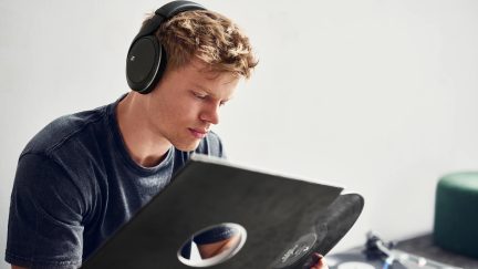 Een man in profiel draagt een zwarte koptelefoon en houdt een zwarte vinylplaat vast in een hoes met een centraal gat.
