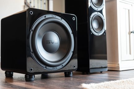 Subwoofer nero lucido con logo SVS, altoparlante per bassi, piedini in gomma, pavimento in legno, diffusore a colonna nero