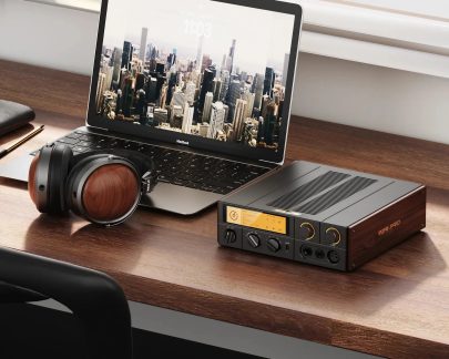 Sort hovedtelefonforstærker med skærm og betjening, placeret nær en MacBook og et par hovedtelefoner på et skrivebord