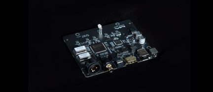 Sort elektronisk printplade i epoxyfiber, set i let skråt trekvartsperspektiv, med integrerede sorte cirkulære XLR-stik, guldbelagt koaksial RCA, guldbelagt HDMI hunstik, samt metalliske USB-A og USB-B porte, sammen med flere chips og grå cylindriske kondensatorer