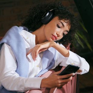 Ung kvinde med sort krøllet hår iført en hvid bluse, lytter til musik med sorte hovedtelefoner på ørerne og lukkede øjne