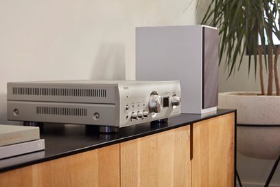 Denon PMA-1700NE sølv hi-fi stereoanlegg, forfra, plassert på et trehylle, med grønn potteplante i bakgrunnen