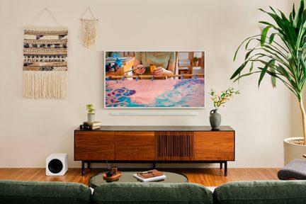 Bílý plochý televizor na stěně s barevným obrazem, pod ním bílá soundbar, dřevěný nábytek a dekorace kolem