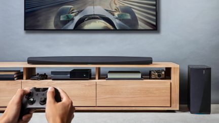 Black soundbar under light wood TV stand, with gaming consoles, speaker, and hands holding controller in front of screen displaying futuristic race car