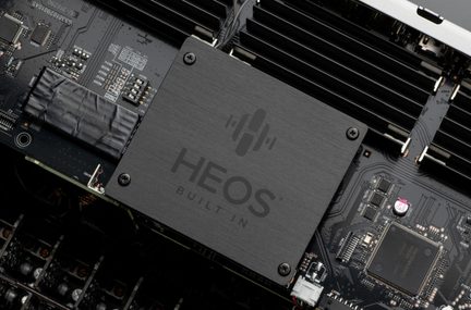 Stambus vaizdas iš viršaus įstrižai į juodą elektroninę plokštę su juodu šlifuoto metalo kvadratiniu dangteliu, pažymėtu „HEOS BUILT IN“, pritvirtintu keturiais varžtais, apsuptu radiatorių su briaunelėmis ir daugybe paviršiuje montuojamų komponentų.