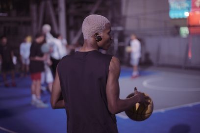 Billedet viser en person bagfra på en travl gade, der holder en brun basketbold. Personen har en sort tanktop på.