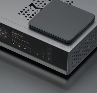 Lecteur audio réseau rectangulaire en aluminium gris clair, vue trois‑quarts avant et dessus, façade écran tactile noir avec commandes affichées, côtés et capot perforés de trous circulaires, petit boîtier carré gris foncé posé sur le dessus
